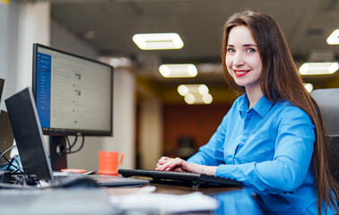 Successful female programmer is sitting at the desk with a computer and works. Beautiful woman looking at camera and smiling in a software company office.