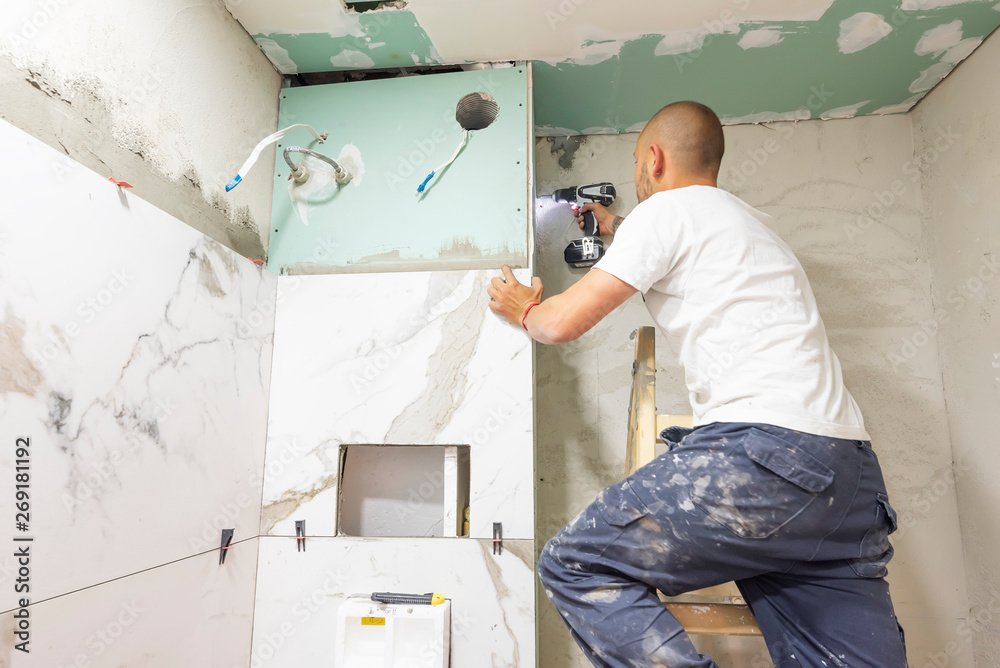 Plumber using electric screwdriver. Bathroom renovation concept. Marble ceramic tiles with spacers and grey cement walls in bathroom. Industrial builder working in toilet - obrazy, fototapety, plakaty 