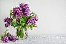 Bouquet Of Purple Lilac Flowers In Glass Vase In White Interior. Space For Text.