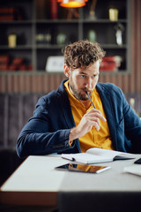 Wall Mural - Young serious bearded Caucasian blogger dressed smart casual writing notes in agenda and looking at laptop while sitting in cafeteria.