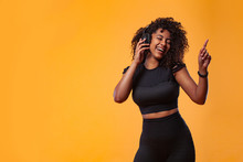 Lifestyle Concept. Portrait Of Beautiful African American Woman With Curly Hair Joyful Listening To Music On Mobile Phone. Yellow Studio Background. Copy Space.