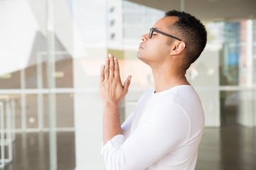 Side view of serious young mixed-race man with closed eyes in white T-shirt, putting hands in praying position. Lifestyle, religion concept