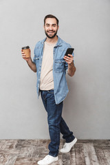 Wall Mural - Full length photo of smiling caucasian man having beard using smartphone and drinking takeaway coffee