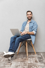 Wall Mural - Photo of casual attractive man having beard typing laptop and using earpods while sitting on chair