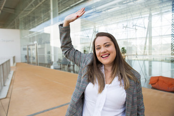 Happy business woman waving with hand outdoors. Lady looking at camera and standing with glass wall in background. Greeting concept. Front view.