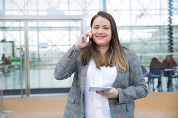 Smiling business woman calling on smartphone outdoors. Lady standing and holding tablet computer with building glass wall in background. Business and technology concept. Front view.