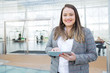 Positive lady with tablet posing in business center. Fat young woman in office jacket holding tablet and smiling at camera. Digital communication concept