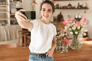 Sticker - Image of brunette young woman taking selfie photo on cellphone and smiling in cozy kitchen