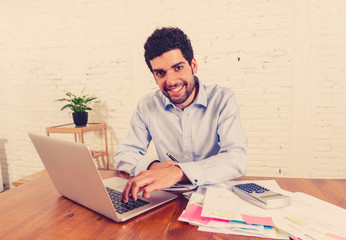 Happy young hipster man sitting on home office desk surrounded by papers calculating and paying bills