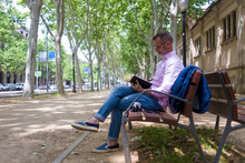 Portrait Of A Bearded Adult Man Sitting On A Wooden Bench In The Park, While Thinking And Writing In A Paper Book