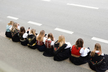 The girls are sitting on the side of the road with mobile phones. A group of people in national Norwegian costumes with smartphones. Concept: traditions and modern technologies, interaction.