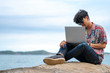© CandyRetriever  - Selective focus young adult man traveler sitting on ocean cliff on tropical island in summer day vacation holding or using laptop and credit card for online shopping or working with ocean backgrounds