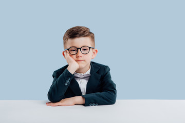 Wall Mural - education. little boy in glasses hold your hand in your head, dressed in school uniform, looking at the camera,  sitting at the white table, isolated on blue background,  Concept of school child