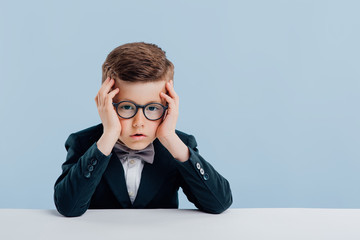 Wall Mural - education. little sad schoolboy holds your hands to the head, looks at the camera, in glasses, sitting at the white table, isolated on blue background, copy space, school concept