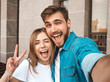 © halayalex - Smiling beautiful girl and her handsome boyfriend in casual summer clothes. Happy family taking selfie self portrait of themselves on smartphone camera. Shows peace sign and winking in the street