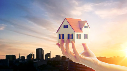 Poster - real estate, accommodation and property concept - close up of hand holding house or home model over sunset sky in tallinn city, estonia background