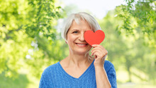 Valentine's Day, Summer And Old People Concept - Portrait Of Smiling Senior Woman Covering One Eye With Red Heart Over Green Natural Background