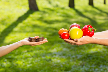 Female and male hands, holding and comparing cookie vs vegetables and fruits. Background of the green park