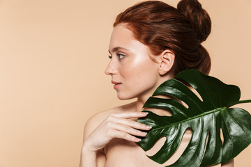 Wall Mural - Pretty young redhead woman posing isolated over beige wall background with leaf green flowers.