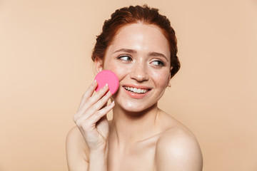 Wall Mural - young redhead woman posing isolated over beige wall background holding makeup sponge
