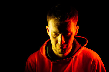 Wall Mural - An adult guy stands with his head tilted down in an orange hooded sweatshirt highlighted in red and yellow on the sides with a pensive and regretful view on a black isolated background.