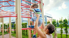 Portriat Of Young Mother Supporting And Holding Her 3 Years Old Child Son On Metal Ladder At Children Playground In Park