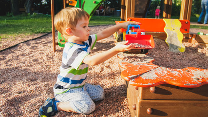 Portrait of cheerful smiling little boy pouring sand in toy truck with trailer. Kids playing and having on playground at park