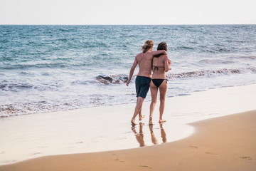 Romantic young people couple walking hugged on the shore with bikini and swimwear for summer vacation lifestyle concept - sea background and youth viewed from back enjoying relationship