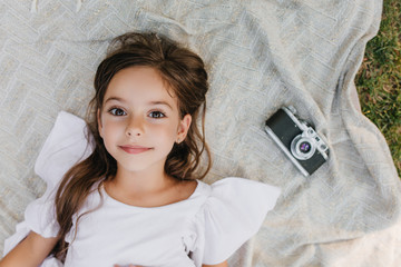 Wall Mural - Inspired little lady with big brown eyes lying on blanket in garden and looking up with gently smile. Overhead portrait of dark-haired girl in white dress relaxing on the ground near camera.