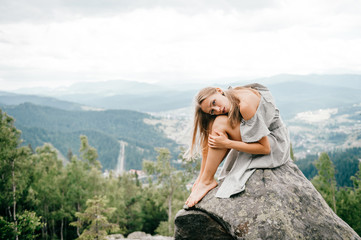 Lonely young girl sits at stone on top of mountain with far view at hills and cloudy sky and hugs her legs with head lying on knees. Beautiful blonde woman outdoor summer  portrait. Female at nature.