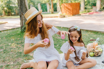 Poster - Barefooted woman in hat with white ribbon sitting on blanket near daughter and eating cookies smiling. Outdoor portrait of happy family joking and fooling around during picnic.