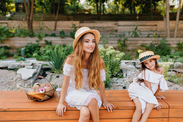 Poster - Joyful woman in lace gown sitting near to picnic basket with legs crossed and looking away. Ecstatic little dark-haired girl in trendy boater chilling on wooden bench with smiling mother.