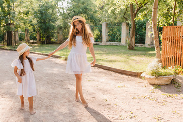 Wall Mural - Pleased slim woman looking with smile at daughter and holding hands with her. Outdoor portrait of enthusiastic young lady walking barefooted in the yard by fence with bushes.