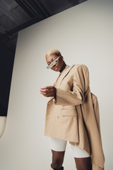 Wall Mural - fashionable african american girl posing in sunglasses and beige jacket on grey