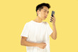 © master1305 - Korean young man's half-length portrait on yellow studio background. Male model in white shirt. Using smartphone. Concept of human emotions, facial expression. Front view. Trendy colors.