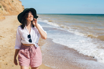 Wall Mural - Stylish hipster girl in hat walking on beach and smiling. Summer vacation. Happy young boho woman relaxing and enjoying sunny warm day at tropical island and blue sky. Space for text