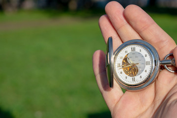 Vintage pocket watch in male hand on a background of green grass. Steampunk watch. Sunny summer day. The clock mechanism is partially visible