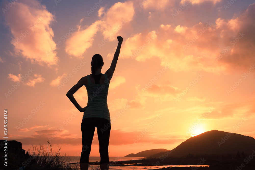silhouette of young strong victorious woman with fist in the air at sunset - obrazy, fototapety, plakaty 