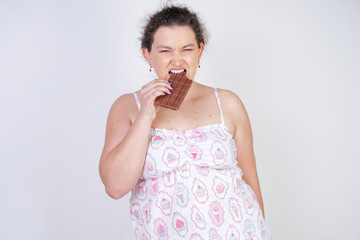 Wall Mural - curvy woman in funny pajamas with a chocolate bar stands on a white background in the Studio