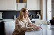 © Ulia Koltyrina - Beautiful young woman in a cozy sweater sitting in the kitchen and listening to music using wireless headphones.