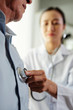 © DragonImages - Close-up of senior man standing while female doctor listening to his heart at the office
