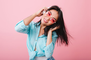 Wonderful asian girl with happy face expression waving hair while listening music. Cute hispanic female model chilling on pink background with favorite song.