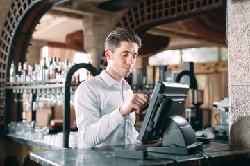 small business, people and service concept - happy man or waiter in apron at counter with cashbox working at bar or coffee shop.