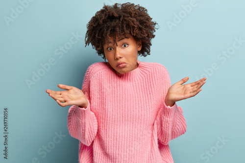 Doubtful African American woman spreads hands, feels uncertain, faces unfamiliar topic, feels perplexed while tries to find answer, wears pink oversized jumper, holds hands sidewards, has no idea