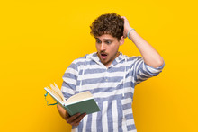 Blonde Man Over Yellow Wall Surprised While Enjoying Reading A Book
