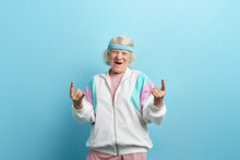 Cute Hipster Grandmother Smiling And Making Rock Sign Against Blue Studio Background