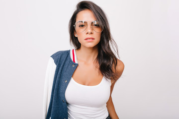 Wall Mural - Close-up portrait of stunning dark-haired girl in white tank-top looking with interest to camera. Indoor photo of adorable european lady in stylish glasses chilling on photoshoot.