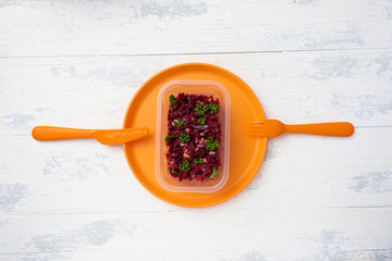 a portion of vinaigrette in a plastic box on a plastic orange plate with a plastic knife and fork on a wooden white table. the view from the top