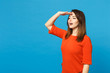 © ViDi Studio - Beautiful young woman wearing orange dress holding hand at forehead looking far away distance isolated over blue wall background, studio portrait. People lifestyle fashion concept. Mock up copy space.