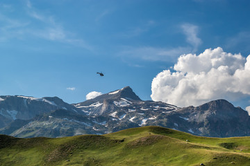 Over a green alpine pasture in a blue sky with white clouds a small black helicopter is flying, French Alps, National Park Vanoise. 